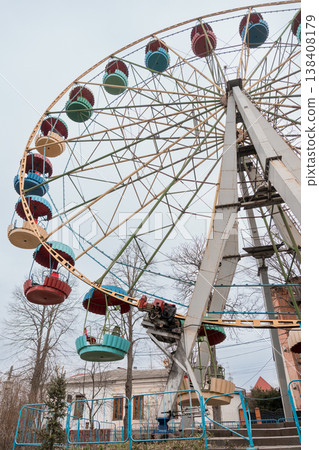 Abandoned Ferris Wheel With Faded Colors Standing Still In Overgrown Park Amidst Bare Trees And Gray Sky Background Showing Rusty Metal Texture Abandoned Ferris Wheel With Faded Colors Standing Still In Overgrown Park Amidst Bare Trees And Gray Sky Background Showing Rusty Metal Texture 138408179