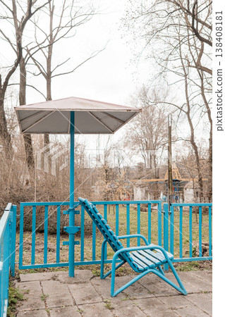 Vintage Blue Patio Chair Under Umbrella on Concrete Terrace Amidst Bare Trees in Overcast Outdoor Setting Vintage Blue Patio Chair Under Umbrella on Concrete Terrace Amidst Bare Trees in Overcast Outdoor Setting 138408181