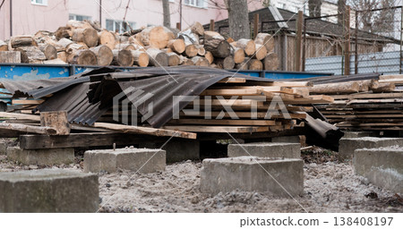 Outdoor Construction Site Featuring Stacked Logs and Planks Covered by Dark Tarps Amidst Concrete Blocks and Snowy Ground with Pink Building Background Outdoor Construction Site Featuring Stacked Logs and Planks Covered by Dark Tarps Amidst Concrete Blocks and Snowy Ground with Pink Building Background 138408197