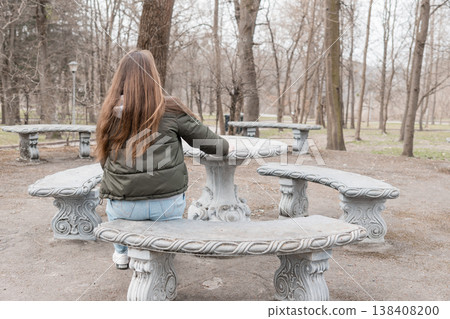 Young Woman With Long Brown Hair Sitting Alone On Ornate Stone Bench In Quiet Park During Overcast Early Spring Day Young Woman With Long Brown Hair Sitting Alone On Ornate Stone Bench In Quiet Park During Overcast Early Spring Day 138408200