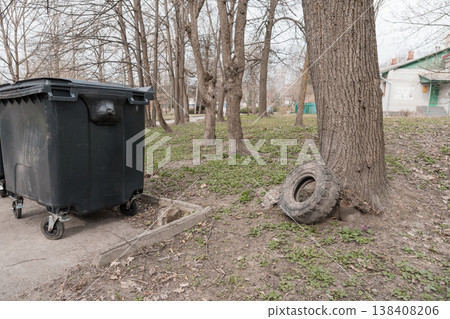 Abandoned Tire Leaning Against Tree Near Black Trash Bin In Urban Park With Bare Trees And Green Grass Under Overcast Sky Abandoned Tire Leaning Against Tree Near Black Trash Bin In Urban Park With Bare Trees And Green Grass Under Overcast Sky 138408206