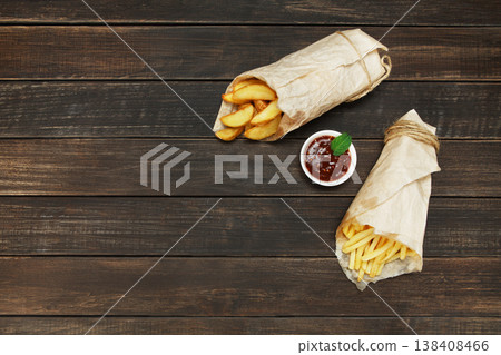 Potato wedges and french fries wrapped in brown wrapping paper. Fast food take away at rustic wood background. Choice of fried potatoes with tomato sauce. Chips, potato slices. Top view, copyspace Potato wedges and french fries wrapped in brown wrapping paper. Fast food take away at rustic wood background. Choice of fried potatoes with tomato sauce. Chips, potato slices. Top view, copyspace 138408466