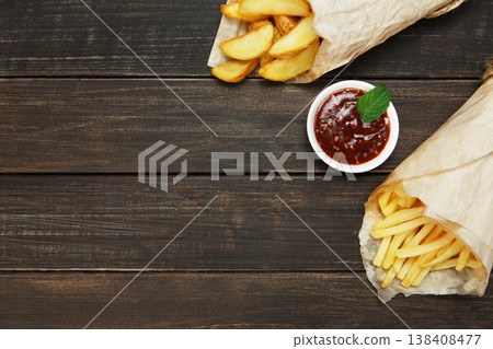 Potato wedges and french fries wrapped in brown wrapping paper. Fast food take away at rustic wood background. Choice of fried potatoes with tomato sauce. Chips, potato slices. Top view, copyspace Potato wedges and french fries wrapped in brown wrapping paper. Fast food take away at rustic wood background. Choice of fried potatoes with tomato sauce. Chips, potato slices. Top view, copyspace 138408477