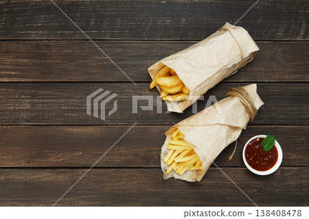 Potato wedges and french fries wrapped in brown wrapping paper. Fast food take away at rustic wood background. Choice of fried potatoes with tomato sauce. Chips, potato slices. Top view, copyspace Potato wedges and french fries wrapped in brown wrapping paper. Fast food take away at rustic wood background. Choice of fried potatoes with tomato sauce. Chips, potato slices. Top view, copyspace 138408478