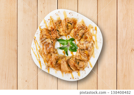 A plate displays neatly arranged dessert rolls topped with a sweet sauce and garnished with mint leaves. The setting features a wooden surface that enhances the food's appearance. 138408482