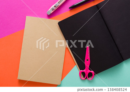 Stationery at modern creative work space. Stylish open notepad with black pages, pink scissors, office knife, marker and craft envelope on abstract multicolored background, top view, copy space Stationery at modern creative work space. Stylish open notepad with black pages, pink scissors, office knife, marker and craft envelope on abstract multicolored background, top view, copy space 138408524