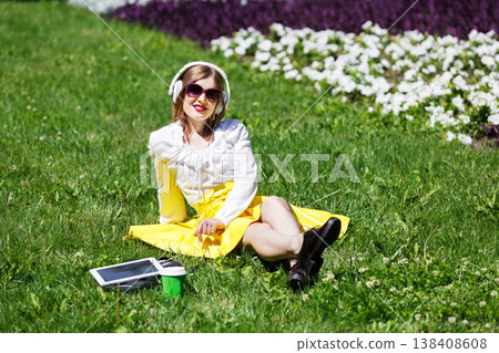 Student girl or young woman listens to the music from tablet pc outdoors. Girl in earphones wears sunglasses sitting on the grass in the park. Music leisure. Having fun, rest, relax in park. Student girl or young woman listens to the music from tablet pc outdoors. Girl in earphones wears sunglasses sitting on the grass in the park. Music leisure. Having fun, rest, relax in park. 138408608