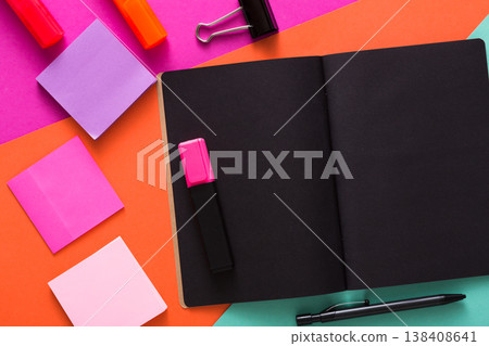 Stationery at modern creative work space. Stylish open notepad with black pages, paperclip, markers and memo notes on abstract multicolored background, top view, copy space Stationery at modern creative work space. Stylish open notepad with black pages, paperclip, markers and memo notes on abstract multicolored background, top view, copy space 138408641