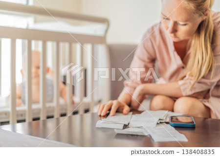 Mother sitting on sofa calculating monthly expenses with receipts while baby lies in crib nearby at home. Stressed mom managing budget and childcare. Financial pressure and parenthood concept Mother sitting on sofa calculating monthly expenses with receipts while baby lies in crib nearby at home. Stressed mom managing budget and childcare. Financial pressure and parenthood concept 138408835