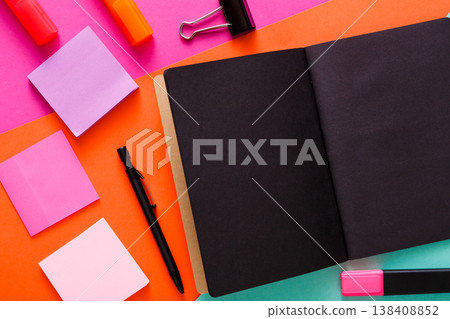 Stationery at modern creative work space. Stylish open notepad with black pages, paperclip, markers and memo notes on abstract multicolored background, top view, copy space Stationery at modern creative work space. Stylish open notepad with black pages, paperclip, markers and memo notes on abstract multicolored background, top view, copy space 138408852