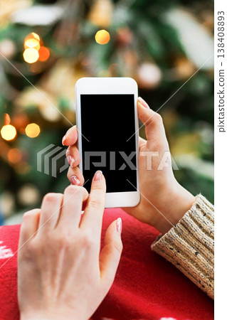 Christmas online shopping. Female buyer makes order at screen of smartphone with copy space. Woman buys presents for xmas eve, sits at home near pine tree with shining lights. Winter holidays sales Christmas online shopping. Female buyer makes order at screen of smartphone with copy space. Woman buys presents for xmas eve, sits at home near pine tree with shining lights. Winter holidays sales 138408893