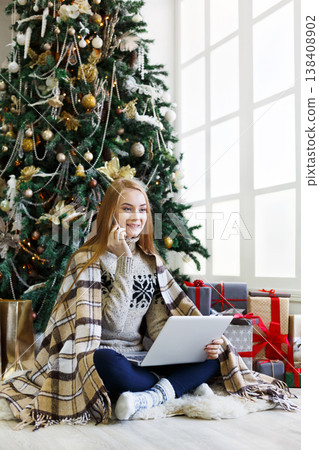 Young woman shopping online on laptop, making order on mobile. Girl sitting under decorated christmas tree among wrapped presents. Preparing to xmas, bying on winter sales. Copy space on window Young woman shopping online on laptop, making order on mobile. Girl sitting under decorated christmas tree among wrapped presents. Preparing to xmas, bying on winter sales. Copy space on window 138408902