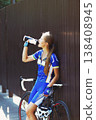 Female sportsman cyclist drinks water from the bottle at the race. Riding racing bicycle. Break, stop at road. Woman cycling on countryside summer road or highway. Triathlon or cycling competition. 138408945