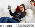 A girl sits on a sofa, focused on a tablet with glasses on. A boy is peeking from behind her, both in a warm indoor space with simple decor. 138408950