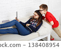 Two kids are sitting on a couch in a bright room. One child lounges on her back, looking at a tablet. The other child stands close, watching the screen with interest. 138408954