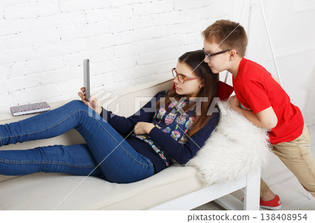 Two kids are sitting on a couch in a bright room. One child lounges on her back, looking at a tablet. The other child stands close, watching the screen with interest. Two kids are sitting on a couch in a bright room. One child lounges on her back, looking at a tablet. The other child stands close, watching the screen with interest. 138408954