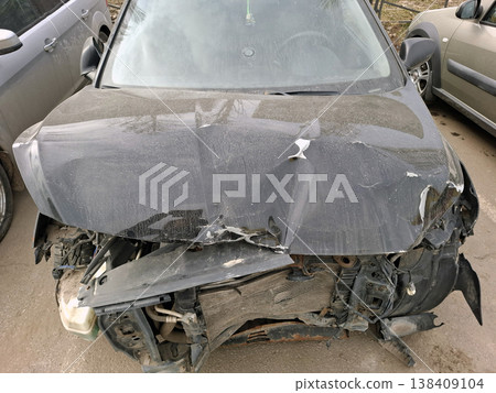 A black car is damaged from a collision. The hood is crumpled, the radiator is smashed, and engine parts are exposed. This is seen at a roadside with other vehicles nearby. A black car is damaged from a collision. The hood is crumpled, the radiator is smashed, and engine parts are exposed. This is seen at a roadside with other vehicles nearby. 138409104