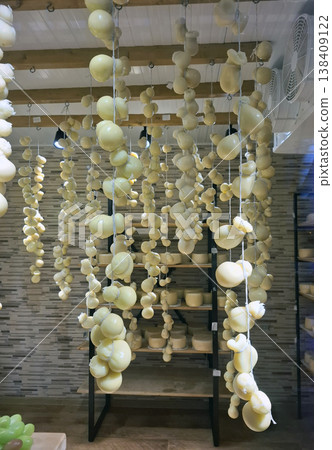 A variety of caciocavallo and scamorza cheeses hang from strings in a rustic cellar in Italy. The room has wooden beams and features a shelf with more cheeses. A variety of caciocavallo and scamorza cheeses hang from strings in a rustic cellar in Italy. The room has wooden beams and features a shelf with more cheeses. 138409122
