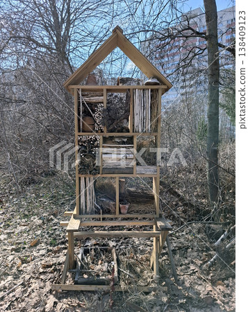 The large insect hotel made of wood is filled with hollow reeds, logs, and straw. It is set in a sunlit park area, providing a home for bugs and supporting local wildlife. 138409123