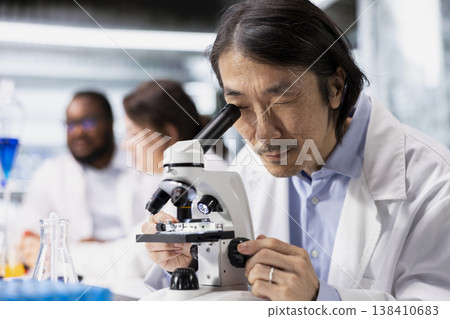 Microscopist operating compound microscope, doing detailed sample observation in controlled laboratory environment. Molecular biologist studies tissue under optical device in biotechnology lab 138410683