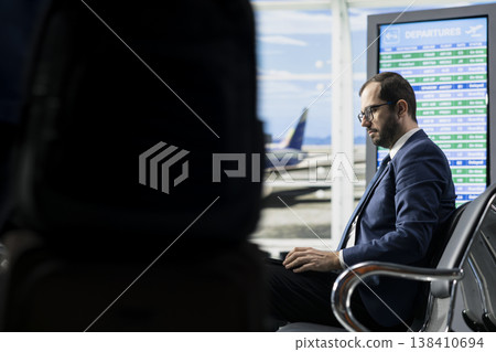 Businessman in airport terminal waiting area works on laptop before departure, frequent flyer corporate travel. CEO traveler near gate with luggage ready, international flight business travel. Businessman in airport terminal waiting area works on laptop before departure, frequent flyer corporate travel. CEO traveler near gate with luggage ready, international flight business travel. 138410694