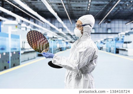 Man in bunny suit evaluating silicon wafer in brightly lit manufacturing plant cleanroom. Engineer checking semiconductor substrate nanometer circuit in modern foundry facility Man in bunny suit evaluating silicon wafer in brightly lit manufacturing plant cleanroom. Engineer checking semiconductor substrate nanometer circuit in modern foundry facility 138410724
