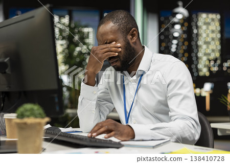 African american drained man experiencing weariness after hours at desk, falling asleep and rubbing his eyes. Overwhelmed anxious and fatigued worker deals with burnout and mental haze. 138410728