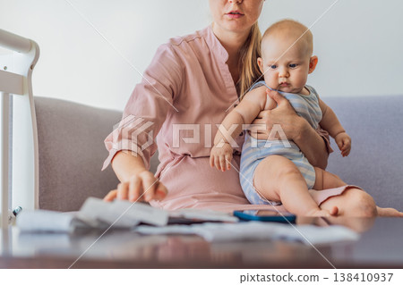 Mother sitting on sofa holding baby while calculating monthly expenses with receipts at home. Stressed mom managing budget and childcare. Financial stress, parenthood and real life concept Mother sitting on sofa holding baby while calculating monthly expenses with receipts at home. Stressed mom managing budget and childcare. Financial stress, parenthood and real life concept 138410937