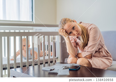 Mother sitting on sofa calculating monthly expenses with receipts while baby lies in crib nearby at home. Stressed mom managing budget and childcare. Financial pressure and parenthood concept Mother sitting on sofa calculating monthly expenses with receipts while baby lies in crib nearby at home. Stressed mom managing budget and childcare. Financial pressure and parenthood concept 138410938
