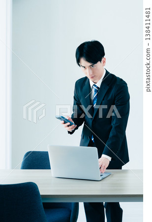 一位年輕的商人正在辦公室裡，一邊拿著智慧型手機一邊操作電腦。 138411481
