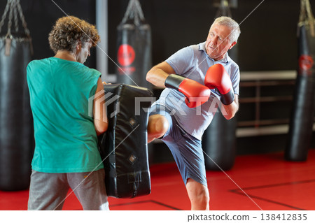 Mature man practicing foot strikes on kick shield with trainer 138412835