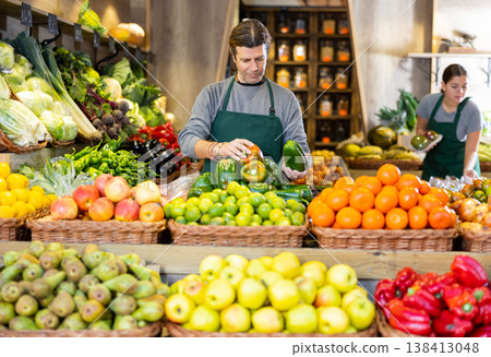 Male seller holding green bell peppers standing in fruit and vegetable section of supermarket 138413048