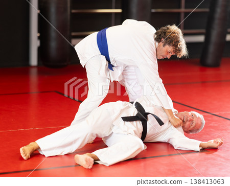 Concentrated blue-belt middle-aged aikido fighter in white gi honing fighting techniques during training kumite with older male partner in dojo Concentrated blue-belt middle-aged aikido fighter in white gi honing fighting techniques during training kumite with older male partner in dojo 138413063