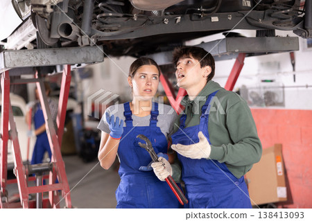 Two young mechanics discussing car repairs under lifted vehicle 138413093