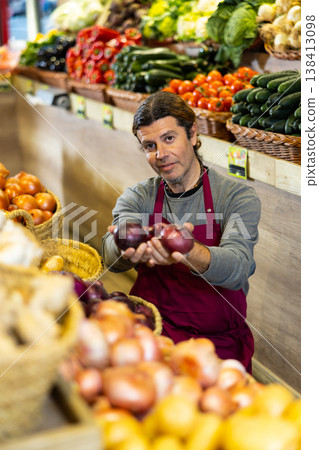 Male seller holding red onion standing in fruit and vegetable section of supermarket Male seller holding red onion standing in fruit and vegetable section of supermarket 138413098