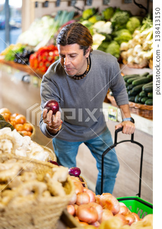 Man shopper examines and chooses red onion on grocery store shelves Man shopper examines and chooses red onion on grocery store shelves 138413150