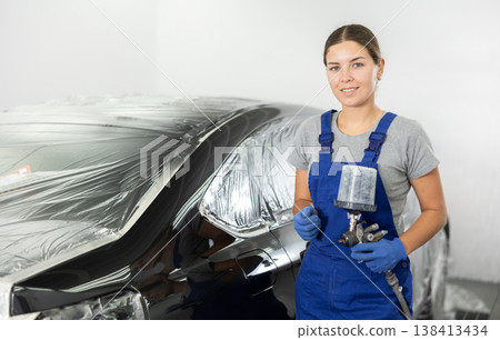 Car painting and automobile repair service. Woman in blue overalls paints car with airbrush pulverizer in paint chamber 138413434