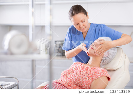 Elderly woman lies in chair during appointment with cosmetologist and enjoys facial massage Elderly woman lies in chair during appointment with cosmetologist and enjoys facial massage 138413539