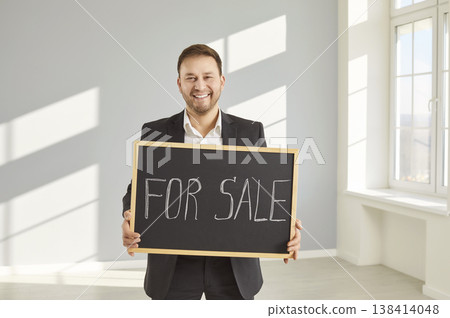 Smiling realtor in suit holding For Sale sign, standing in bright empty room with large windows. Smiling realtor in suit holding For Sale sign, standing in bright empty room with large windows. 138414048