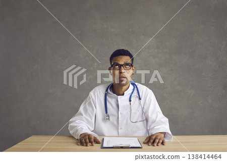 Young black doctor sitting at office desk, looking up and thinking about something Young black doctor sitting at office desk, looking up and thinking about something 138414464