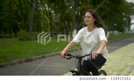 Young woman in white t shirt and black leggings riding bicycle in park front view. 4k 138415161