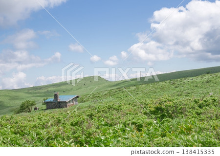 A lush green plain and a mountain lodge, a blue sky and white clouds—a summer scene. A lush green plain and a mountain lodge, a blue sky and white clouds—a summer scene. 138415335