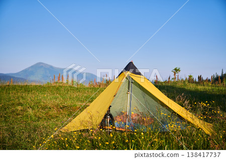 Yellow tourist tent on grassy meadow, surrounded by wildflowers, with majestic mountains in background. Clear blue sky and sunlight create vibrant and peaceful camping scene in nature. Yellow tourist tent on grassy meadow, surrounded by wildflowers, with majestic mountains in background. Clear blue sky and sunlight create vibrant and peaceful camping scene in nature. 138417737