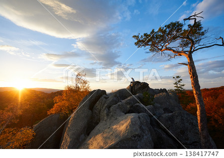 Sun sets, casting golden glow over rocky landscape and autumnal trees. Lone person atop rocks, silhouetted against vibrant sky. Dovbush Rocks, Carpathian mountains, Ukraine. Sun sets, casting golden glow over rocky landscape and autumnal trees. Lone person atop rocks, silhouetted against vibrant sky. Dovbush Rocks, Carpathian mountains, Ukraine. 138417747