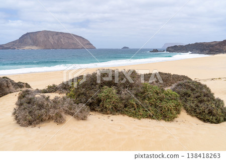 A tranquil beach scene in La Graciosa, Lanzarote, featuring golden sand, ocean, and distant volcanic hills under a cloudy sky. A tranquil beach scene in La Graciosa, Lanzarote, featuring golden sand, ocean, and distant volcanic hills under a cloudy sky. 138418263