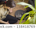 Woman Closely Checks Robot Vacuum Among Houseplants. Casual Homeowner With Ponytail Assesses Robot Vacuum While Tending Indoor Plants In Warm Sunlight 138418769