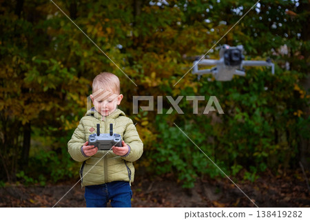 A young child stands in a lush park surrounded by vibrant autumn leaves, skillfully maneuvering a drone with a remote control. Excitement fills the air. 138419282