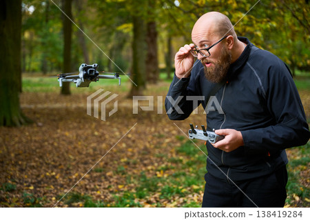 A man focuses on maneuvering his drone through the vibrant autumn landscape. Golden leaves cover the ground as he adjusts his glasses, showcasing a moment of concentration and joy. A man focuses on maneuvering his drone through the vibrant autumn landscape. Golden leaves cover the ground as he adjusts his glasses, showcasing a moment of concentration and joy. 138419284