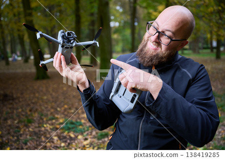 A man with a beard smiles and points at his drone in a colorful forest. Yellow and orange leaves surround him, creating a joyful autumn scene. A man with a beard smiles and points at his drone in a colorful forest. Yellow and orange leaves surround him, creating a joyful autumn scene. 138419285