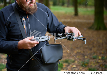 A man with a beard stands in a peaceful park, getting ready to launch his drone. The colorful autumn leaves surround him, creating a tranquil atmosphere. 138419289